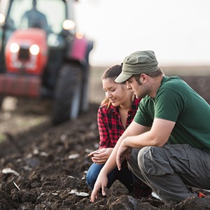 young couple kneeling in a field examining the soil near farm equipment